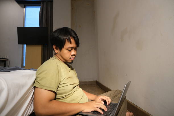 mężczyzna z azji południowo-wschodniej opierający się o łóżko ze złą postawą i piszący na laptopie - southeast asian ethnicity men laptop image type zdjęcia i obrazy z banku zdjęć