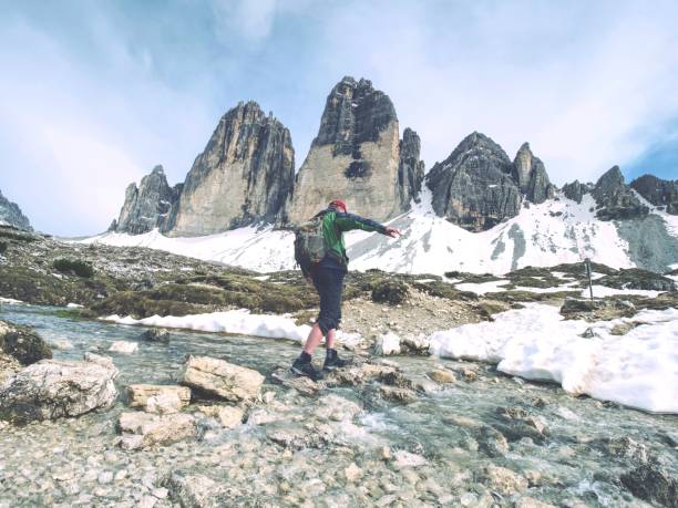 Hiker Man Jumping Over Stream At Tre Cime Di Lavaredo Massif. Tour Around Sharp Peaks, Dolomite Alps