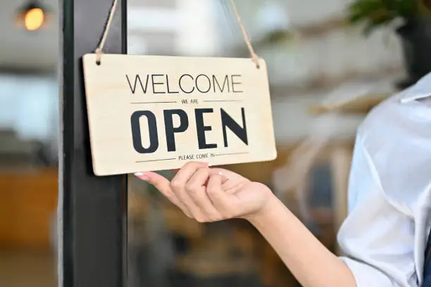 Photo of Female barista holding a coffee shop welcome sign in front of the coffee shop.