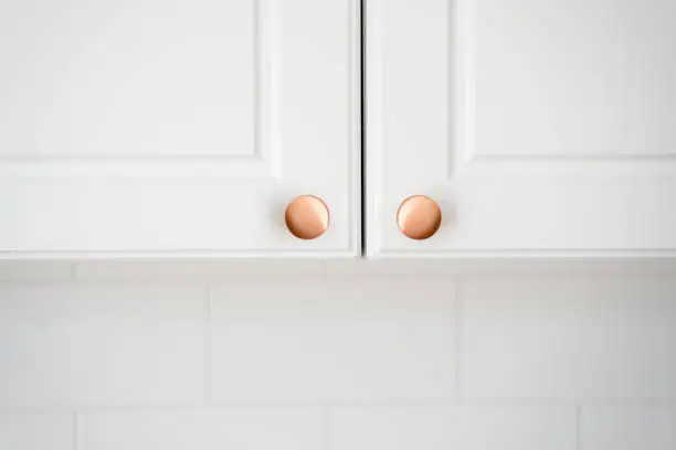 Concept of replacement or install of new handle on furniture in classic style. Close up view of two small round copper door knob on white kitchen cabinet over copy space tiled wall