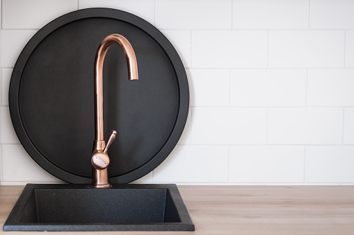 New copper faucet installed over empty sink in the modern kitchen interior. Black and metal breakfast tray standing behind water tap, against white copy space tiles on the wall. Household concept