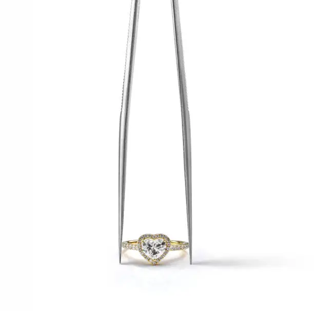 Heart shaped yellow gold diamond halo engagement ring on surface, held between vertical pair of diamond sorting gemstone tweezers.