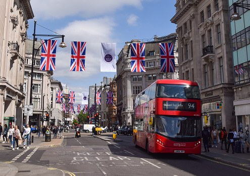 London, UK - May, 2023. People crossing street and sit on steps of monument at Piccadilly Circus Street in London during daytime in springtime with Piccadilly Circus Shopping center behind and huge advertisement board. British Flag and Coronation Flag  Landscape format.