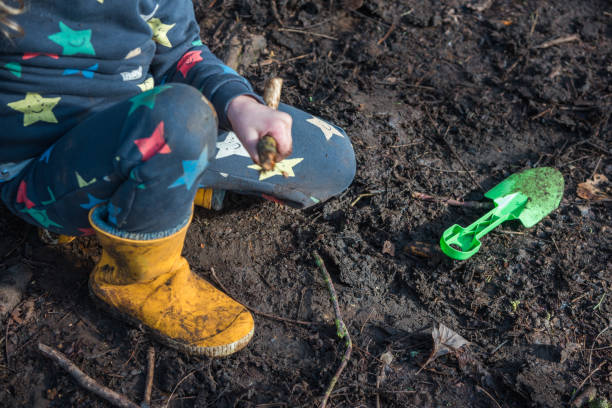 A child is playing with stick and mini shovel on the dirt in a forest. Only appearing the body of the child kneeling on the soil, wearing dark blue clothes with colorful stars and yellow boots.
