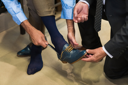 Low section of mature male customer wearing shoe with shoehorn by owner kneeling at showroom