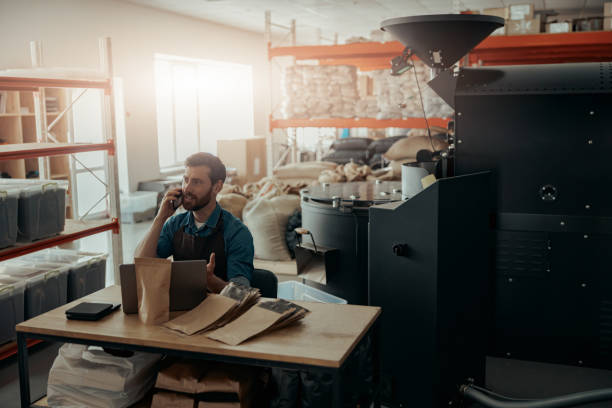 dueño de un negocio de una pequeña fábrica de tostado de café hablando por teléfono en su lugar de trabajo - roasted machine bean mixing fotografías e imágenes de stock