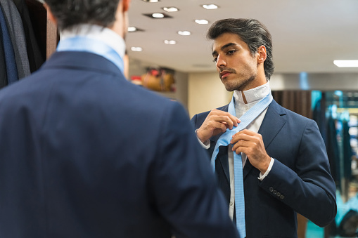 Handsome mid adult man looking at mirror reflection while wearing necktie in clothing store