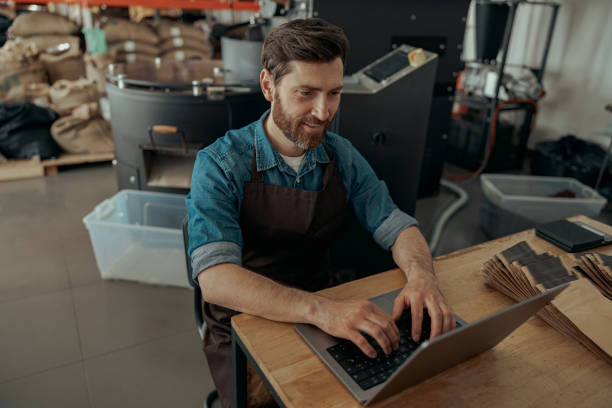 propietario de un negocio de una pequeña fábrica de tostado de café que trabaja en su lugar de trabajo con una computadora portátil - roasted machine bean mixing fotografías e imágenes de stock