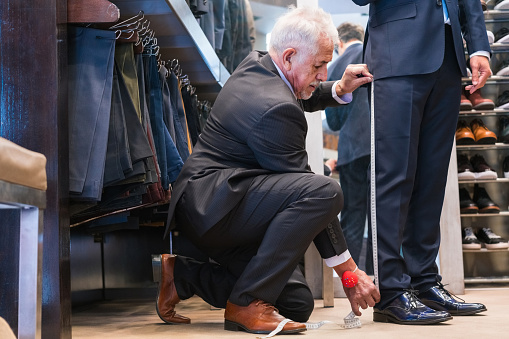 Senior male tailor measuring customer's length of pant while kneeling at clothing store