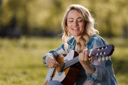 Happy woman sitting on the maedow playing guitar and singing during spring day