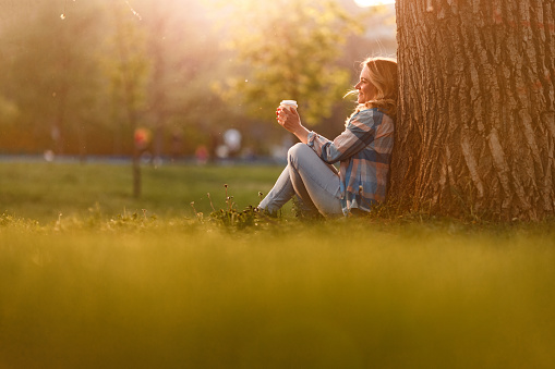 Smiling woman leaning on the tree in nature and holding cup of coffee