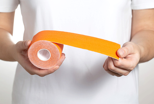 The girl holds a roll of orange kinesiology tape in her hands on the background of a white T-shirt.