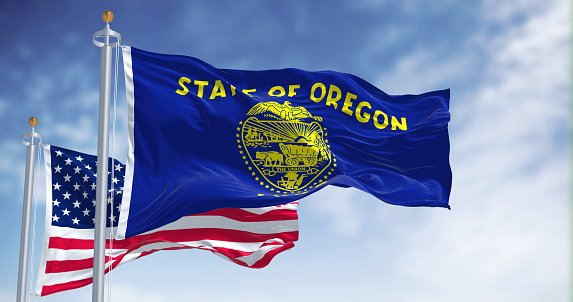 The Oregon state flag waving along with the national flag of the United States of America. In the background there is a clear sky. Oregon is a U.S. state in the northwest