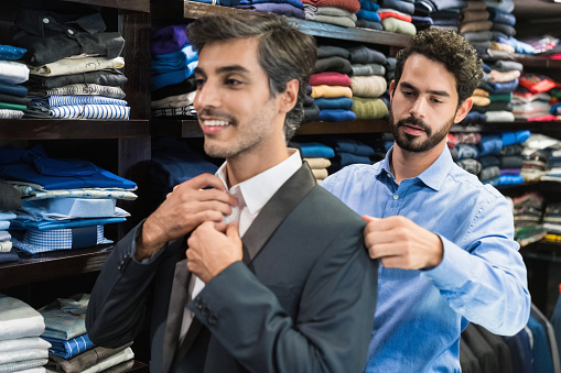 Young owner helping male customer wearing suit at menswear clothing showroom.