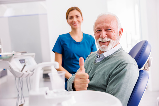 Portrait of satisfied senior male patient sitting at dental chair at dentist office showing thumbs up and smiling with doctor in the background