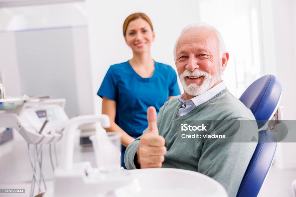 Senior man at dentist office showing thumbs up Portrait of satisfied senior male patient sitting at dental chair at dentist office showing thumbs up and smiling with doctor in the background Dentist Stock Photo