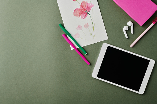 Part of workplace of designer with watercolor drawing, tablet, earphones, small pink notebook, pen and highlighters on grey background