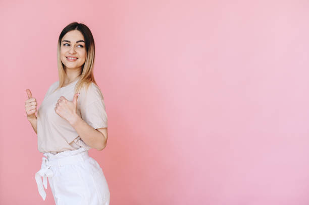 istock A girl posing half-side in light-colored T-shirt shows thumbs up with both hands on pink background 1397889205