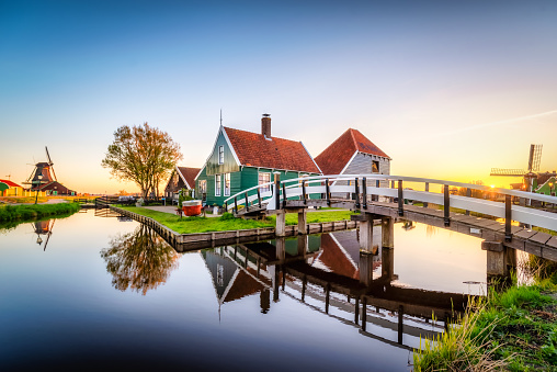 April 17, 2022 - Zaanse Schans, Zaandam, Netherlands: Antique and traditional Dutch cheese farm houses with a wooden bridge across a canal. Reflection in calm water. Sun is rising in the background, Location is Zaanse Schans, one of the popular tourist attractions of The Netherlands.
