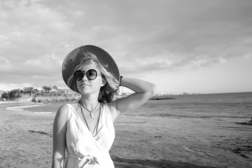 Beautiful boho woman enjoying summer vacation, walking at the beach. Portrait of blonde woman wearing fashionable sunglasses and hat. Tourism. Real people lifestyle.