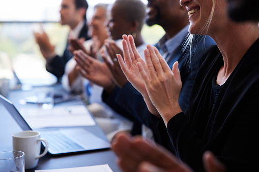 Close-up of a diverse group of smiling businesspeople sitting in a row at a table and clapping after a boardroom presentation