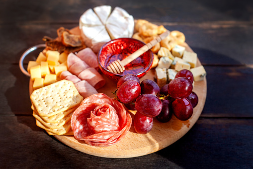 Round cutting board with slicing assorted cheese, sausage rosette, ham, cracker, grapes and figs with honey. Charcuterie and cheese platter on the terrace with sunbeams