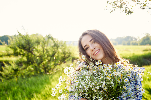 Beautiful Young Woman sitting on the field in green grass and blowing dandelion. Outdoors. Enjoy Nature. Healthy Smiling Girl on summer lawn. Allergy free concept. Gorgeous slim mixed race Caucasian Asian model posing on holidays. Side view