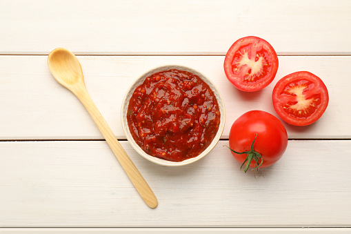 Delicious adjika sauce in bowl with spoon and tomatoes on white wooden table, flat lay