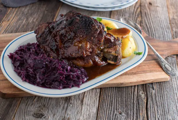 Homemade holiday roast with roasted turkey shank, red cabbage, boiled potatoes and delicious gravy. Served on a plate isolated on wooden table background with copy space