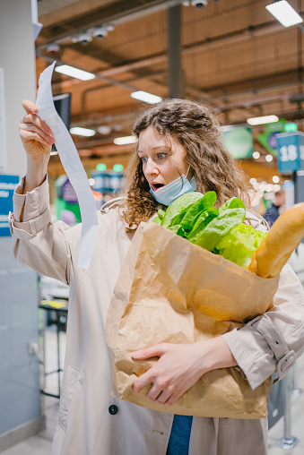 Woman in a medical mask in a supermarket holds a long check and groceries in a paper bag and looks at the high prices in surprise in shock.
