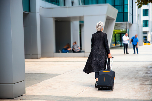 Mature handsome lady pulling her suitcase in the urban city. Back of Older white hair female on heels walking with luggage to airport