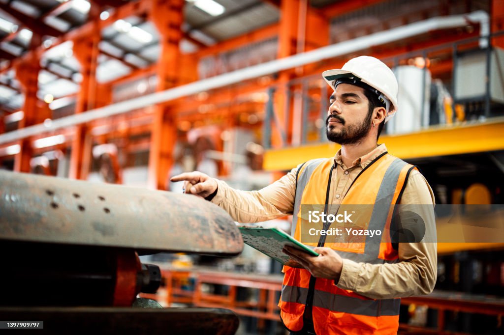 Men industrial engineer wearing a white helmet while standing in a heavy industrial factory behind. The Maintenance looking of working at industrial machinery and check security system setup in fact General View Stock Photo