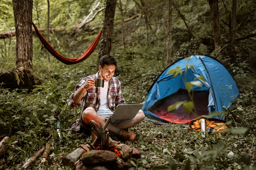 Young handsome man having a coffee while working from his camp in a forest