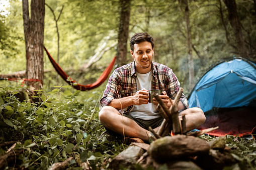 Young casually clothed hiker drinking tea near the campfire