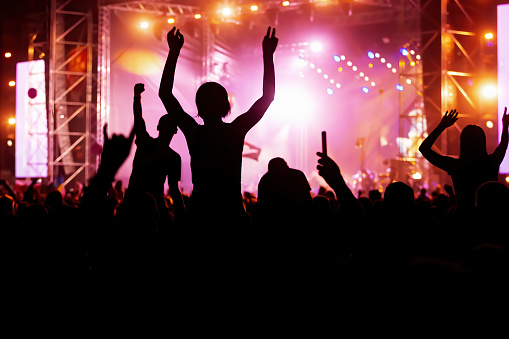 Rear view of large group of people enjoying a concert performance. There are many hands applauding and taping the show. Multi colored lasers and spot lights firing from the stage.\nSilhouettes have been significantly liquified.