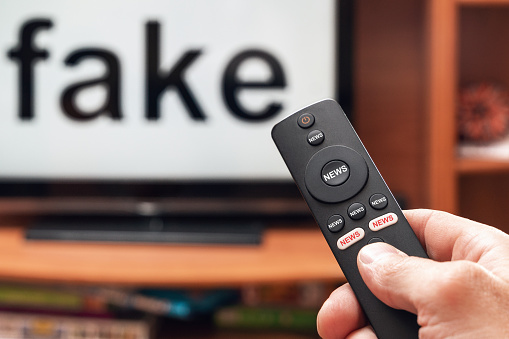 A man's hand holds a TV remote control, all the buttons on the remote control are signed news, on the TV the inscription is fake.