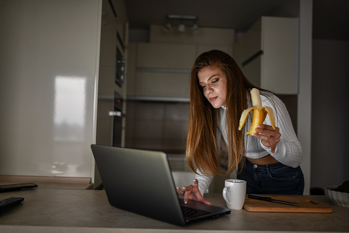 Modern Blond Female Beauty Eating Banana While Working On Laptop In Kitchen