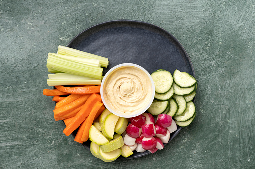 Healthy snack chickpea hummus with various fresh vegetables and cereals on a gray platter. Cucumbers, radishes, celery, carrots, tomatoes, zucchini, red onions, parsley, cilantro. Healthy food, copy space for text