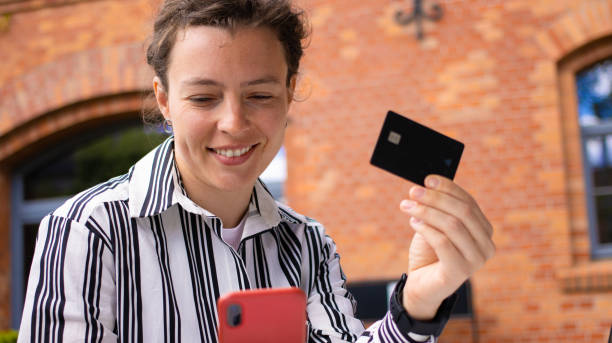 젊은 성인 백인 여성 소비자는 검은 신용 카드를 들고 붉은 벽돌 부디 백그라운드에서 스마트 폰을보고 웃고 있습니다. 온라인 뱅킹 거래, 안전한 모바일 뱅킹 개념 수행 - buiding 뉴스 사진 이미지