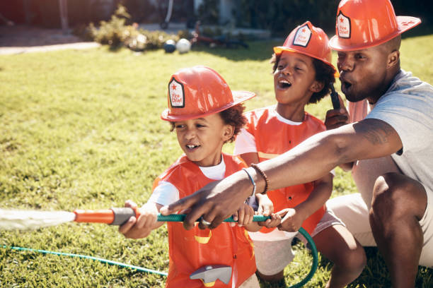 vater spielt mit seinen söhnen draußen. kleine jungen, die als feuerwehrleute verkleidet waren. afroamerikanische jungs spielen draußen. brüder, die mit einem schlauchrohr im garten spielen. geschwister, die wasser aus einem schlauchrohr sprühen. - african ethnicity brother ethnic little boys stock-fotos und bilder