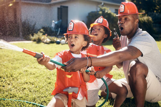 kleine jungen in feuerwehrkostümen. afroamerikanischer vater, der mit seinen kindern spielt. kleine jungen, die im garten spielen. jungen, die wasser aus dem schlauchrohr draußen sprühen. aufgeregte brüder spielen - african ethnicity brother ethnic little boys stock-fotos und bilder