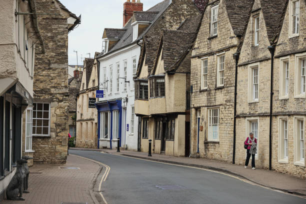 Elderly lone woman wandering around a quaint small street full of historical 16th century town houses in the English Cotswold market town of Cirencester in Gloucestershire stock photo