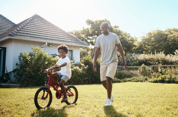 adorable niño afroamericano aprendiendo a andar en bicicleta afuera con su padre. papá e hijo divirtiéndose en su patio trasero en un día soleado - african descent cycling men bicycle fotografías e imágenes de stock