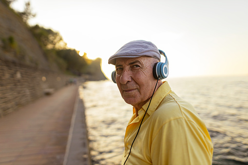 Senior, white man wearing hat, yellow shirt, headphones  and standing beside a beautiful sea. Listening music and enjoying.