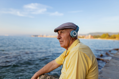 Senior, white man wearing hat, yellow shirt, headphones  and sitting beside a beautiful sea.