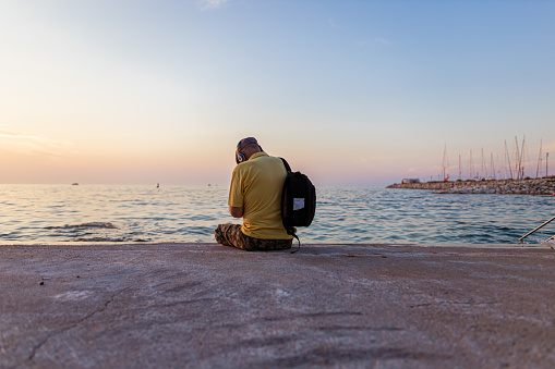 Active senior man wearing hat, yellow shirt, headphones, backpack  and sitting beside a beautiful sea. Listening music and enjoying.