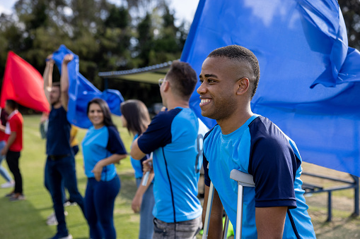 Injured African American soccer player watching the game from the bench while standing in crutches with a group of fans - sports concepts