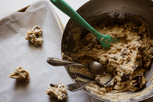 silver mixing bowl of chocolate chip cookie dough with a spatula and two silver spoon sticking out of it. Along side a baking sheet with ball of cookie doughs