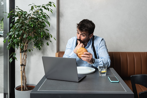 Young successful freelancer business man watching movie on his laptop computer during her lunch break. Exchange student waiting for video call with his family while he eating big fat hamburger