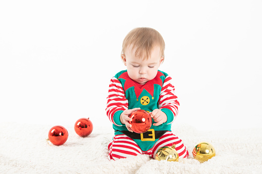 Stock studio photo with white background of a baby in elf costume grabbing christmas balls with serious expression